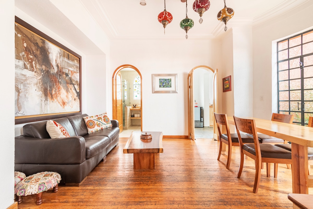 A spacious living area features a dark leather sofa adorned with colorful cushions. A wooden coffee table sits centrally, with a dining table and four chairs arranged nearby. Decorative hanging lamps provide subtle illumination, and arched doorways lead to other rooms.