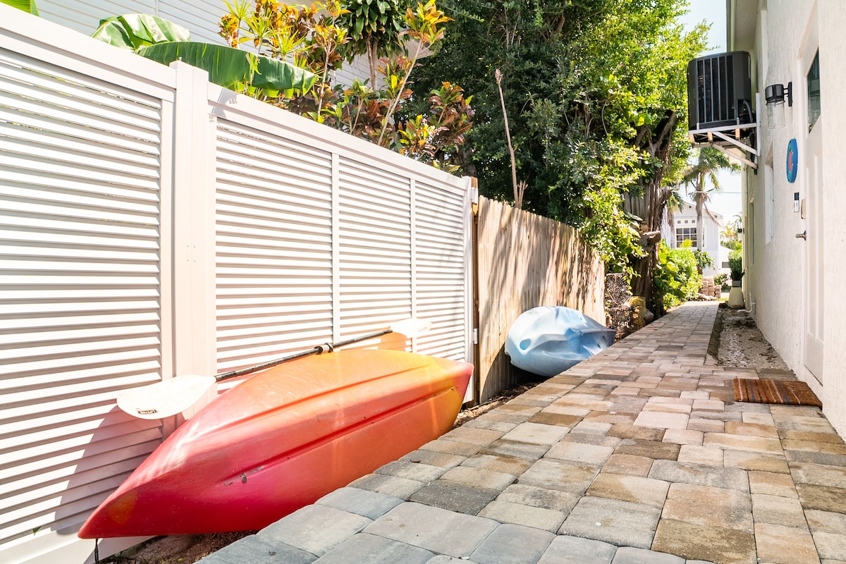 A pathway lined with vibrant tropical vegetation is shown, featuring two kayaks resting against a white fence. One kayak is bright orange, while another is a soft blue, both positioned beside a stone paved walkway leading to the residence.