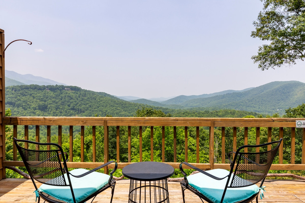 A private deck features two black wire chairs with aqua cushions and a round black table. The railing provides a view of the expansive green mountains and valleys in the distance, enhancing the sense of tranquility and connection to nature.