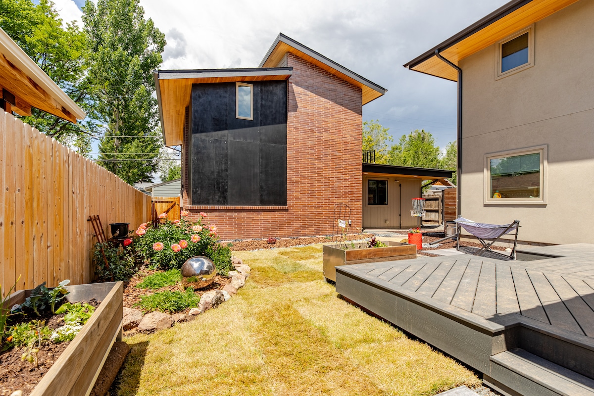 A shared backyard features a blend of grass and landscaped areas, bordered by wooden fencing. The two-story accessory dwelling unit showcases a brick exterior and large windows. A wooden deck offers a space to relax, while garden beds with plants add visual interest to the patio area.