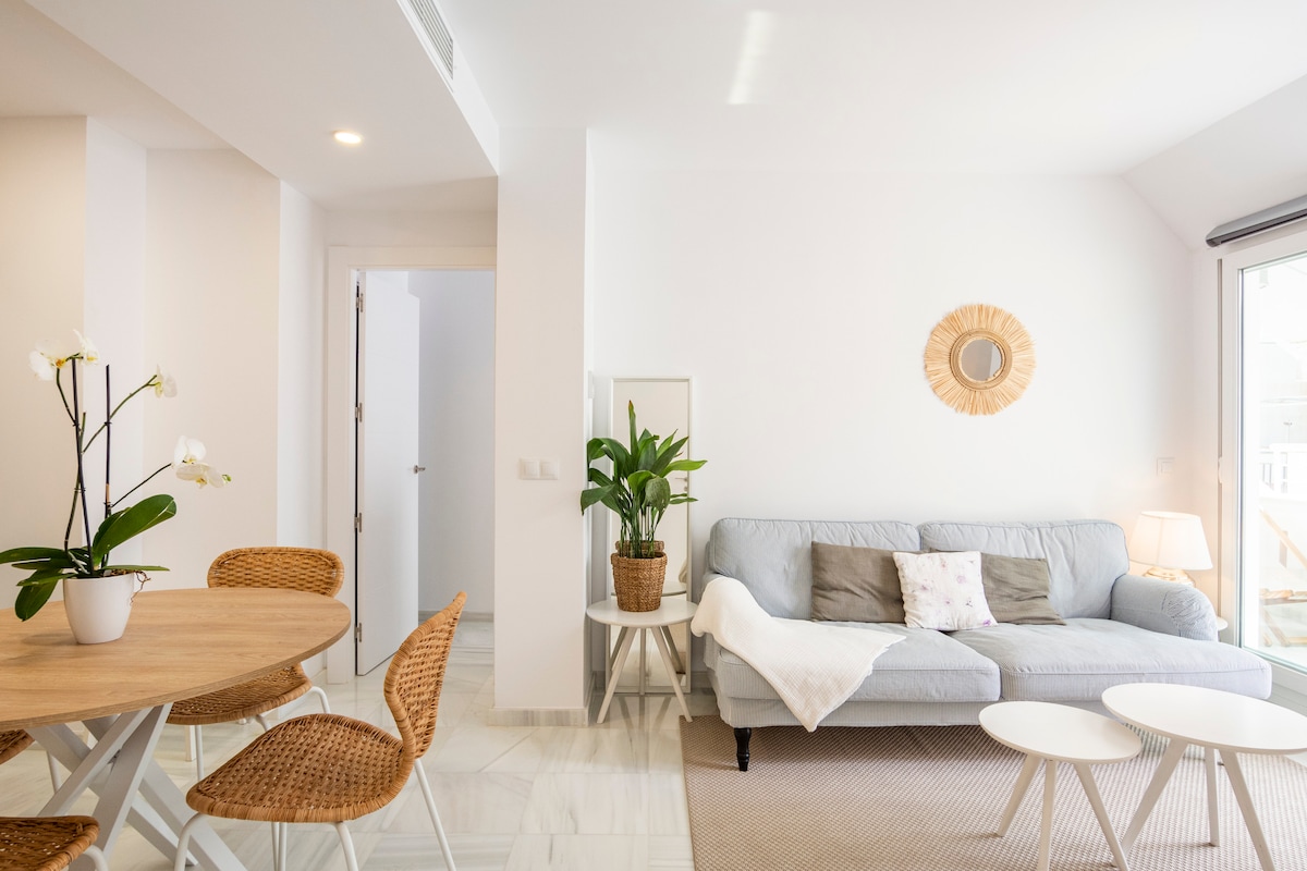 A bright and airy living space features a light gray sofa accented with decorative pillows. A round dining table with four rattan chairs occupies one side, while a potted plant adds a touch of greenery. Large windows allow natural light to fill the room.