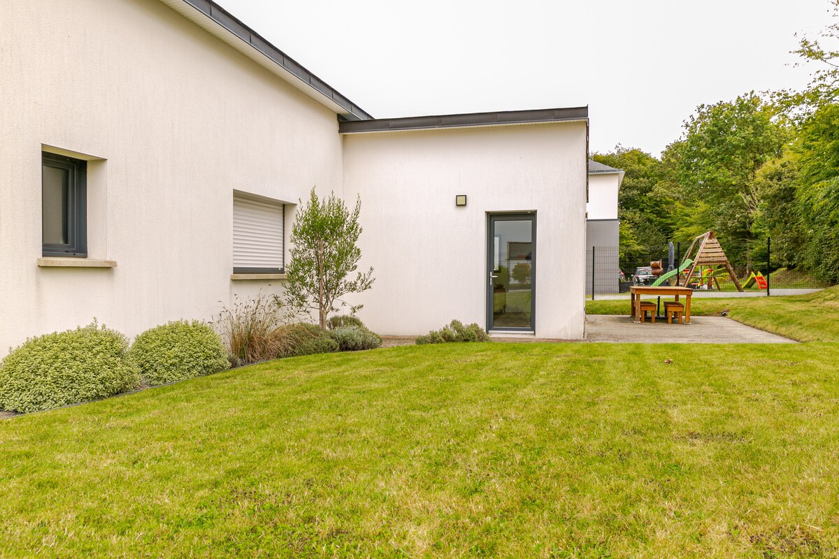 A modern house exterior is visible, featuring a neat lawn surrounded by low shrubs. A sliding glass door leads into the home, with outdoor seating visible nearby. Children’s play equipment can be seen in the background, enhancing the family-friendly environment.