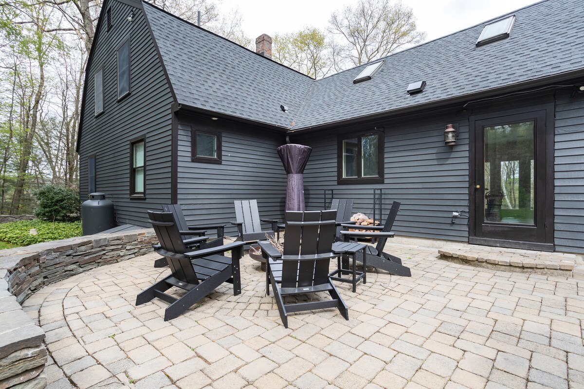 A stone-paved outdoor patio features a circular arrangement of black wooden Adirondack chairs surrounding a central table. An umbrella offers shade, and a nearby black water tank is partially visible. The house's dark exterior and large windows create a cozy atmosphere.