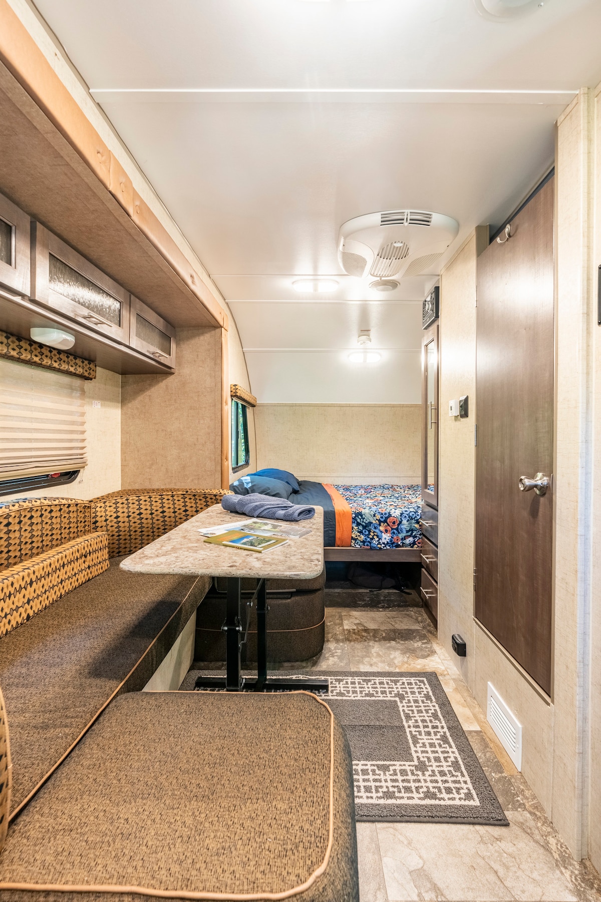 The interior of a small RV features a dinette area with a padded bench and table set against a neutral wall. Opposite is a cozy sleeping space with a colorful quilted bedspread. Light from the ceiling fixtures highlights the compact layout and amenities.