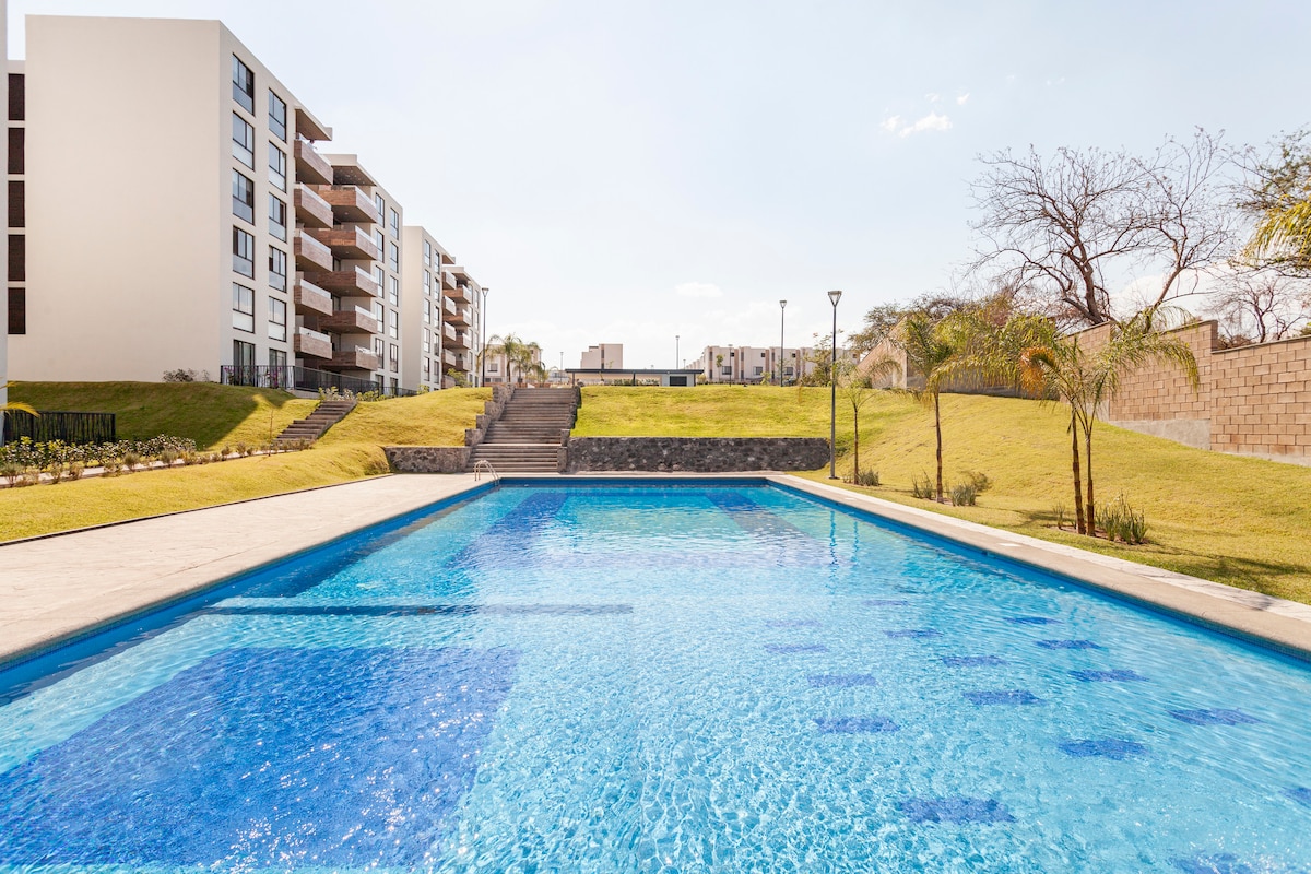 A spacious outdoor pool is framed by well-maintained lawns and modern apartment buildings. Clear blue water reflects the sky and surrounding greenery, while gentle slopes lead to the pool area, offering an inviting space for relaxation and recreation.