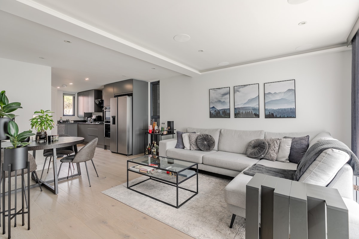 The open-plan living space features a spacious gray sofa with decorative pillows, a glass coffee table, and a round dining table with four chairs. Two mountain-themed artworks are displayed on the wall. The kitchen area with dark cabinetry is visible in the background.