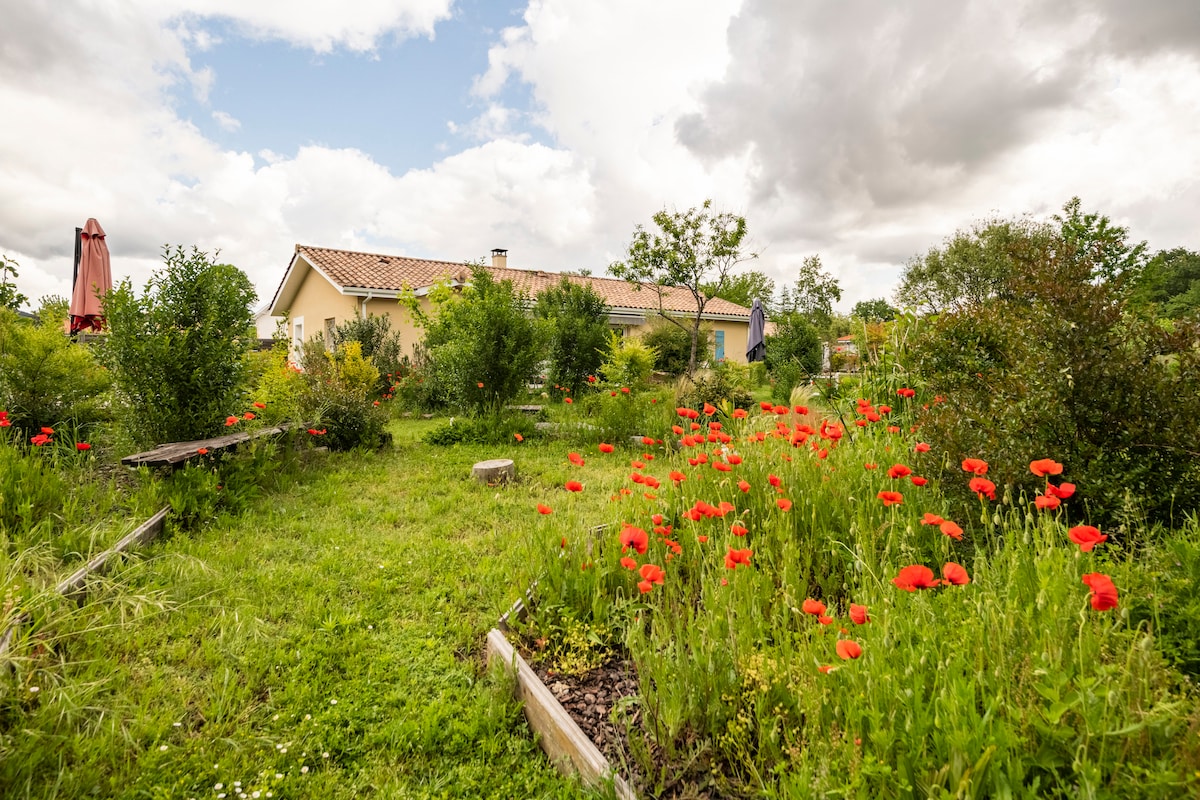 A spacious garden is filled with vibrant red poppies, surrounded by lush greenery. The villa is seen in the background, featuring a sloped roof and natural landscaping. Shady areas are provided by small trees and shrubs, enhancing the tranquil outdoor setting.