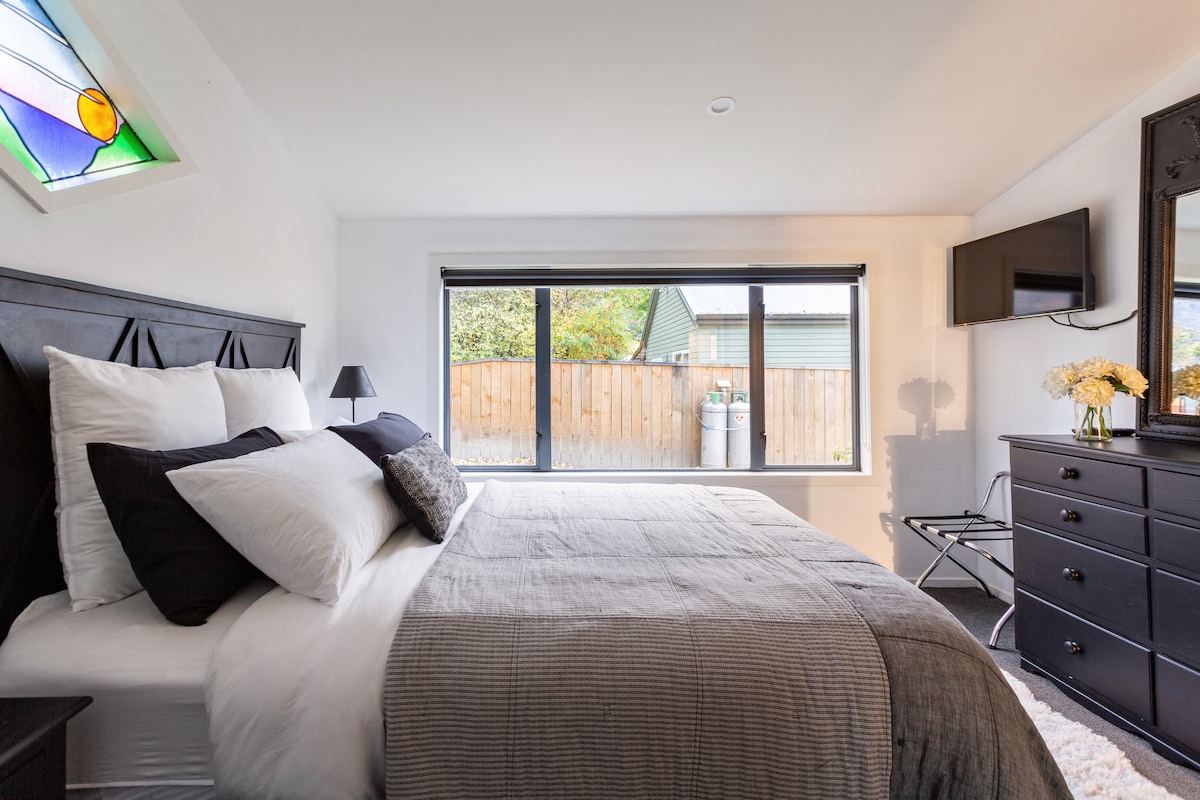 A spacious sleeping area features a large bed with crisp white linens and dark pillows. Natural light enters through wide windows, showcasing a view of the outdoor space. A small lamp and a sleek chair complement the stylish dresser and wall-mounted television.