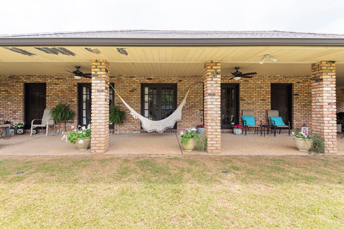 A spacious porch features three brick columns that support the roof. Colorful seating is arranged on either side, with a hammock suspended between two columns. Potted plants add greenery, while light-colored gravel covers the ground, creating an inviting outdoor space.