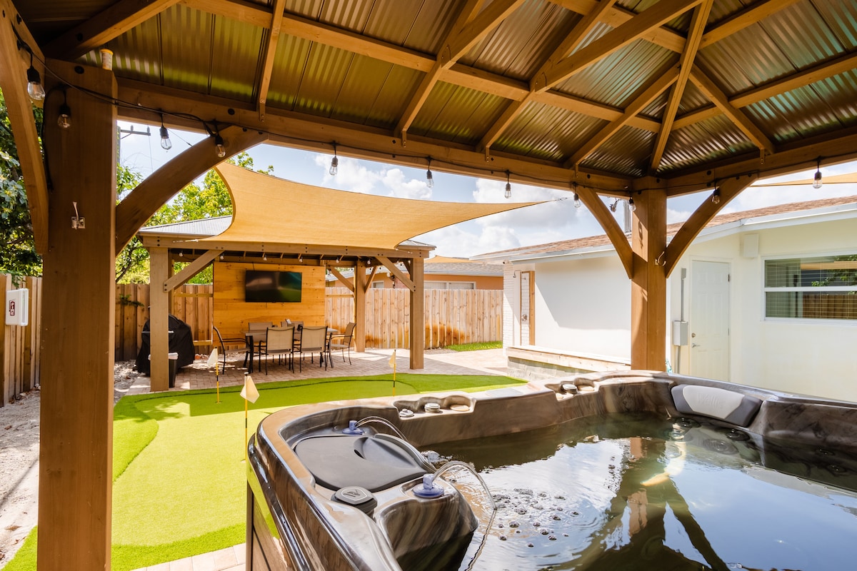 A spacious outdoor area is featured, showcasing a hot tub under a covered structure. String lights are hung overhead, creating a relaxed atmosphere. Nearby, a putting green and a dining table can be seen, providing options for both leisure and meals.