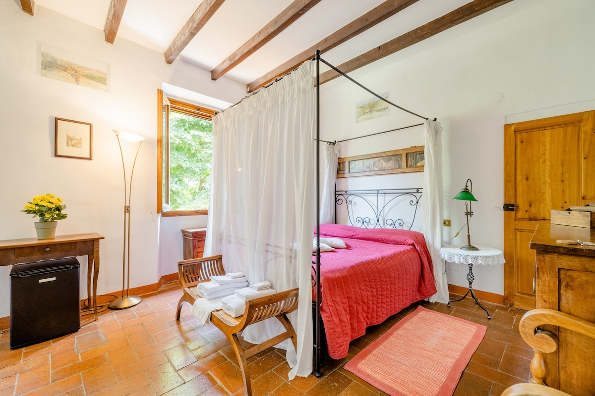 A spacious room features a canopied bed with a vibrant red coverlet. Two wooden chairs are positioned beside a small table. Natural light filters through a large window, complemented by a floor lamp. The room is adorned with wooden beams and light-colored walls.