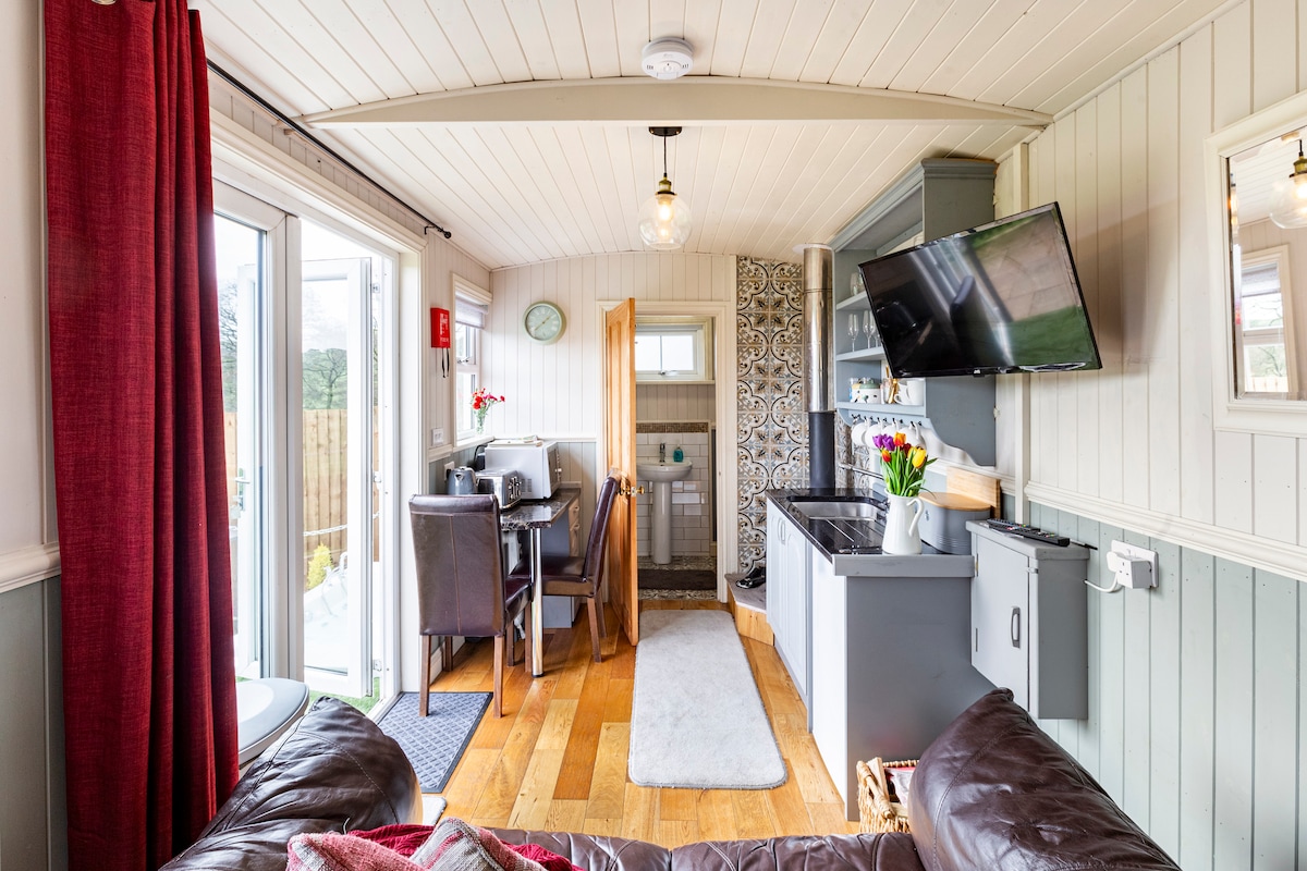 A bright open-plan living area features a comfortable sofa, wooden flooring, and a wall-mounted TV. A dining table and chairs are placed near the kitchen, which includes modern appliances. Natural light enters through double doors leading to an outdoor space.