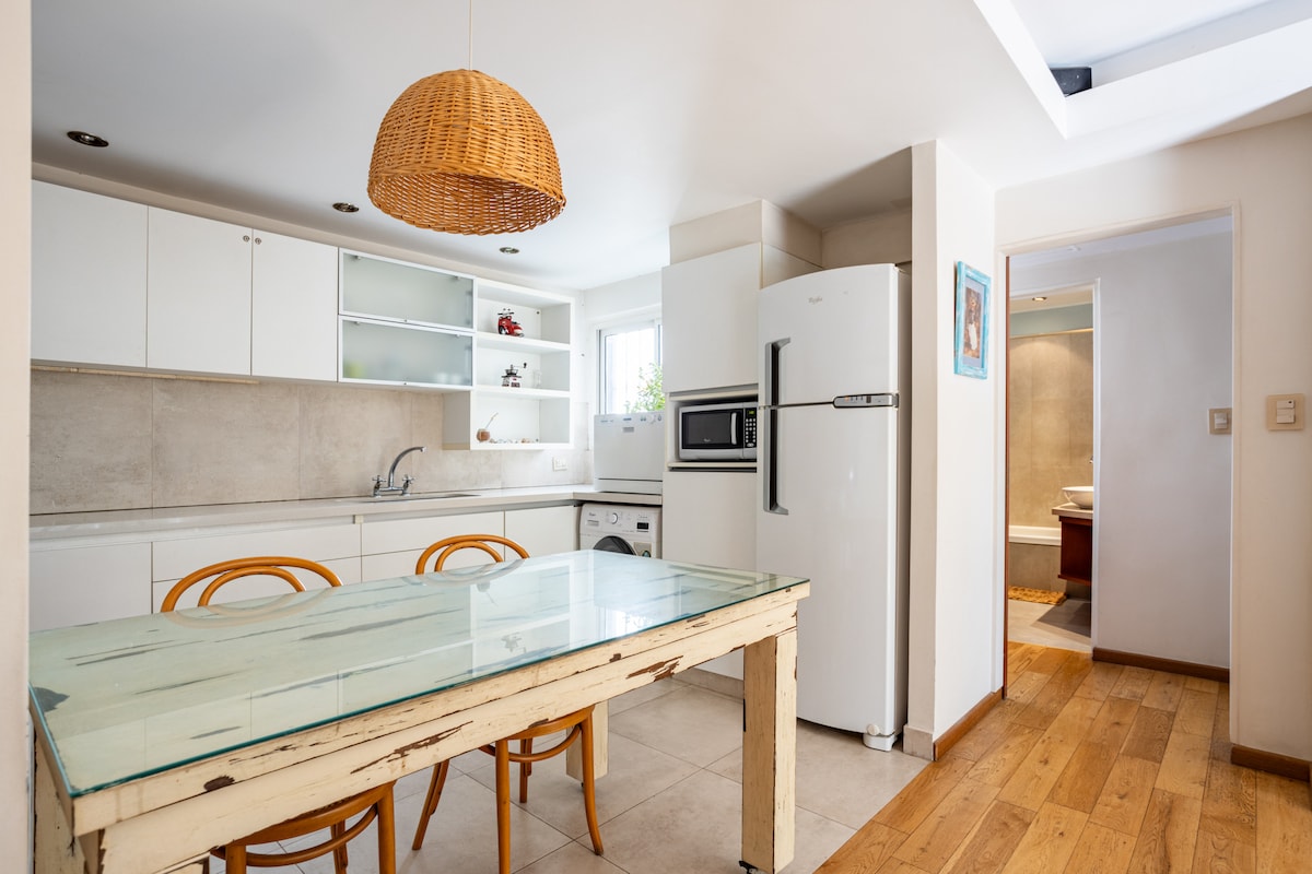 An inviting kitchen area features a wooden table surrounded by curved chairs. Light streams in through a window above the sink, illuminating white cabinetry and modern appliances, including a refrigerator and stove. A woven pendant light adds warmth to the space.