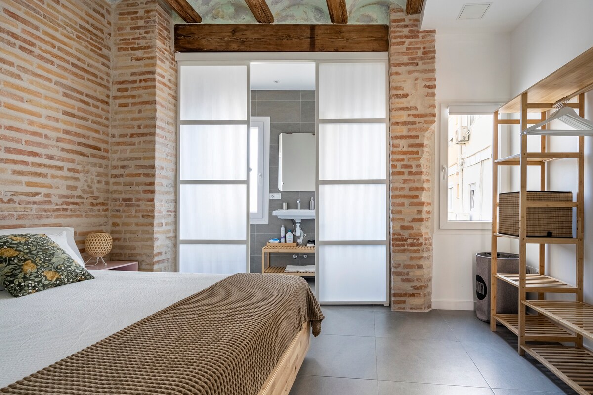 A spacious bedroom is framed by exposed brick walls and wooden beams. The bed is dressed in a neutral coverlet, with a decorative pillow adding a touch of color. Light filters through sliding translucent panels that separate the bedroom from the bathroom, complemented by a window providing natural light.