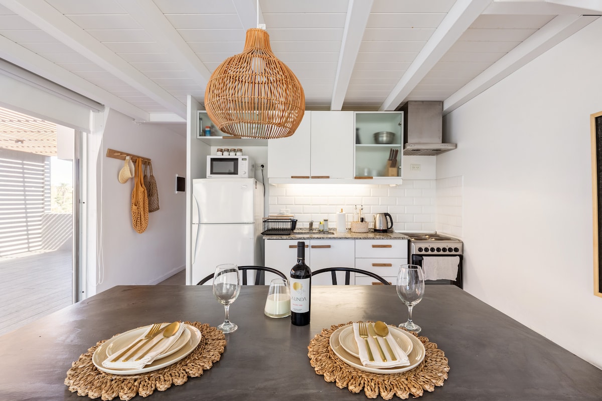 A dining area features a dark table set with elegant dishes and glasses. A bottle of wine is centered on the table, while a woven pendant light hangs overhead. A modern kitchen can be seen in the background, equipped with essential appliances and natural light streaming in.