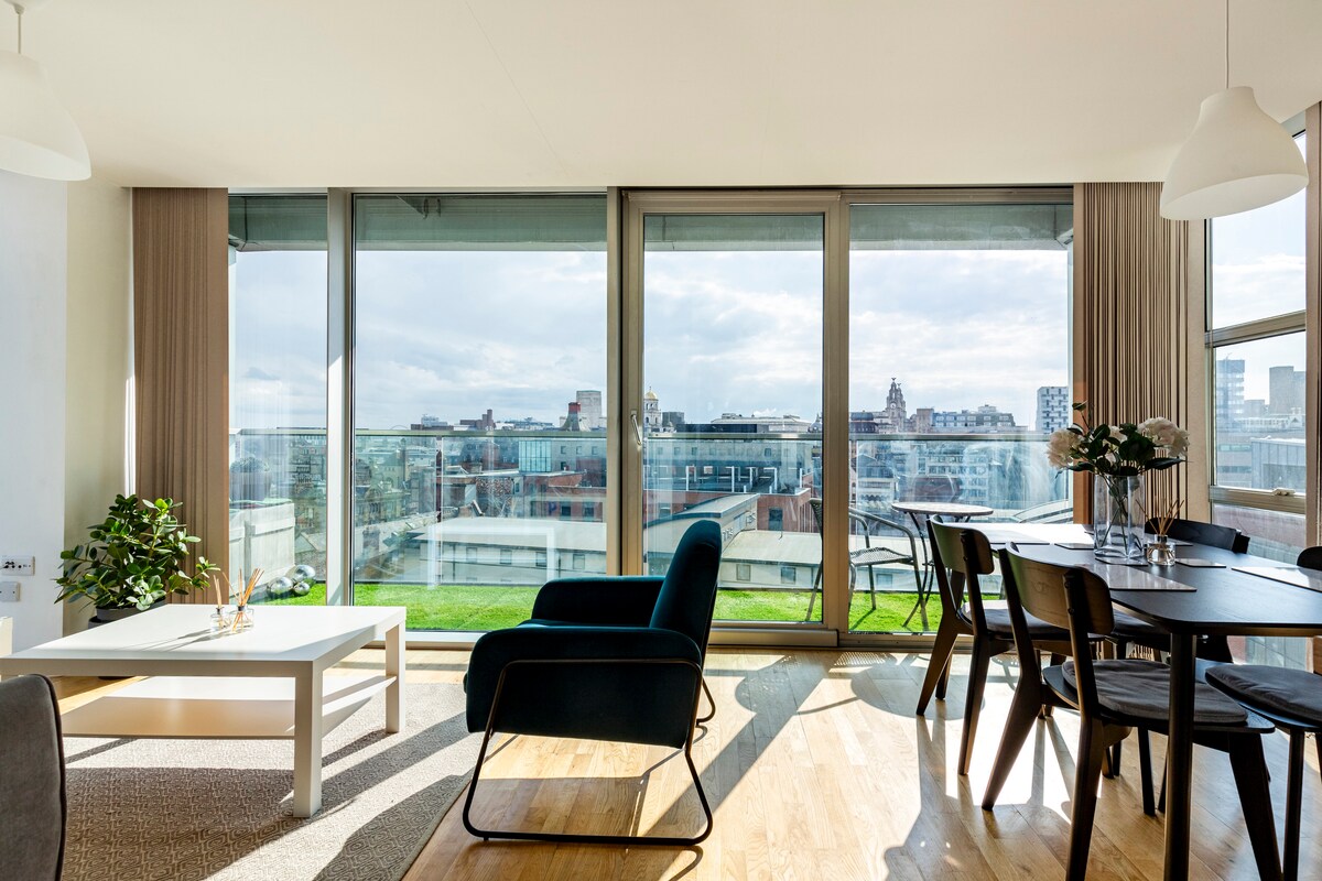 A spacious living area is featured, with large glass windows allowing natural light to flood the space. A blue armchair is positioned beside a white coffee table, while a dining table is set with black chairs. Outside views of the cityscape are visible.