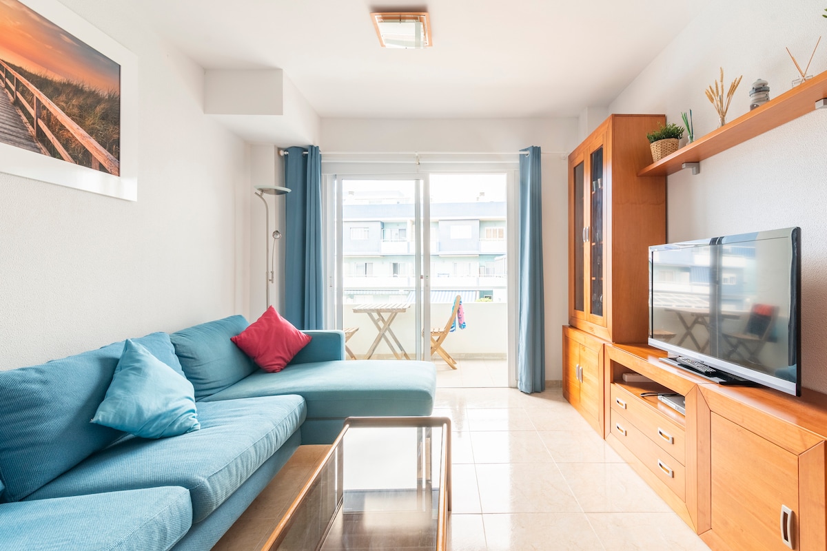 A bright living area is highlighted by a spacious blue sofa adorned with colorful cushions. Natural light floods in through large glass doors, leading to a balcony with a small table and a chair. A wooden entertainment unit displays a television, complemented by light-colored wall shelves.
