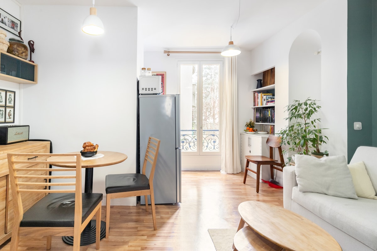 A well-lit living area features a cozy sofa and a small wooden dining table with black chairs. A modern refrigerator is positioned nearby, complemented by shelving displaying books and decorative items. Natural light floods the space through the large window, which is adorned with light curtains.