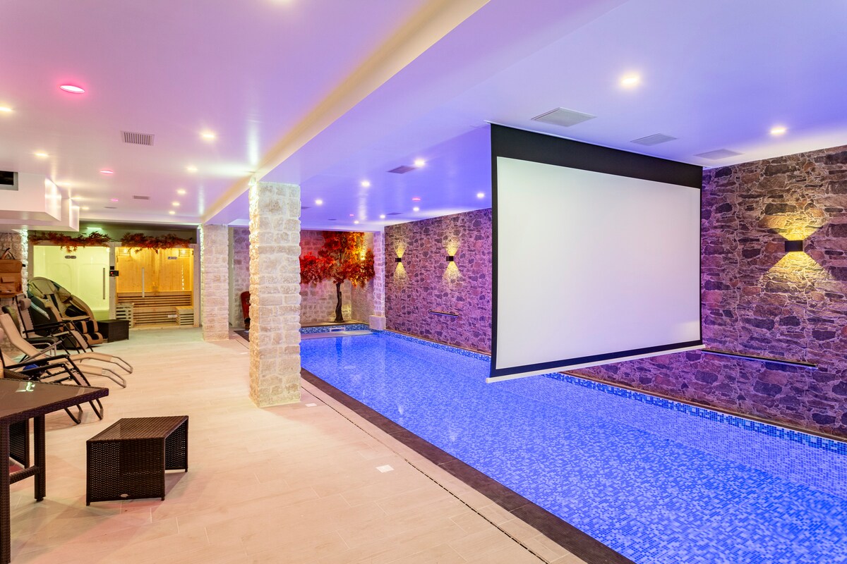 An indoor heated swimming pool features a vibrant blue surface, surrounded by stone walls with soft lighting. A large screen is positioned at one end for cinematic experiences. Lounge chairs are placed nearby, complementing a relaxing atmosphere.