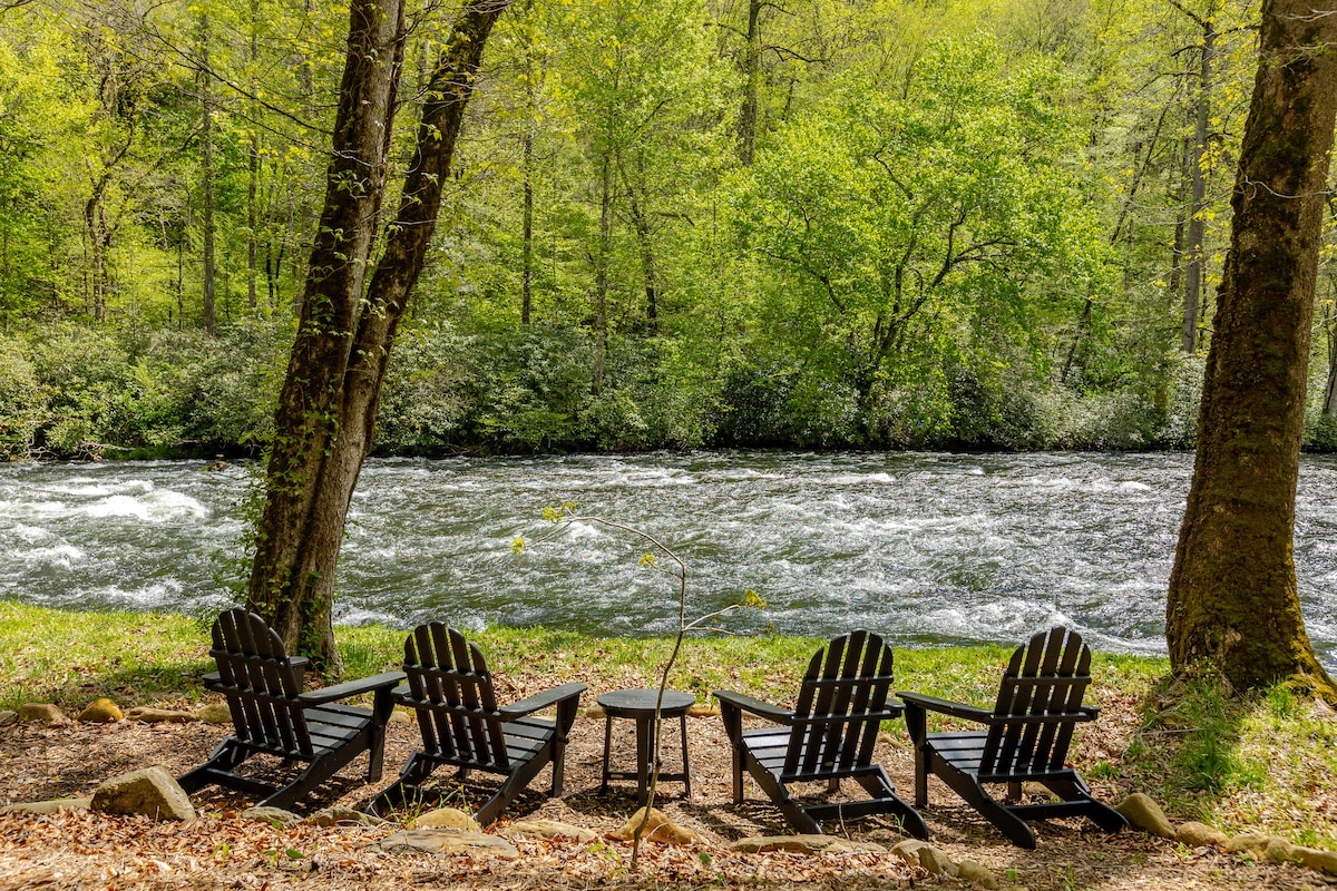 Four Adirondack chairs are arranged around a small table by the riverside, framed by lush green trees. The river flows steadily, with the sound of rushing water evident. This shaded area offers a serene spot for relaxation and enjoying the natural surroundings.