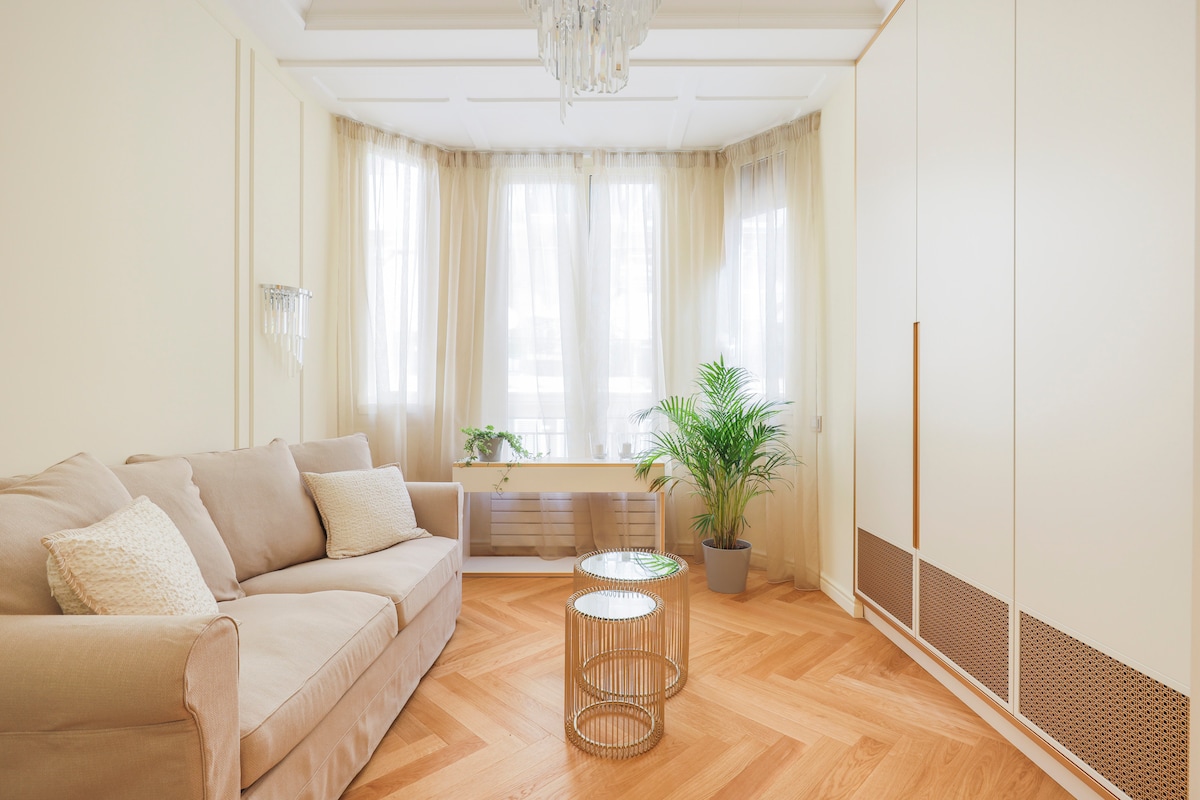 A light-filled living area features a neutral sofa positioned against a window with sheer curtains, allowing for soft natural light. A small, round glass table sits in front, complemented by a decorative plant in a pot, adding a touch of greenery to the space.
