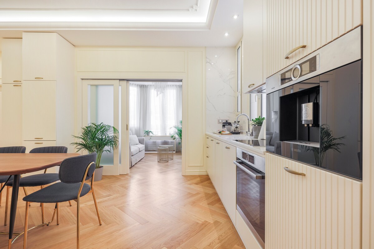 A modern kitchen area features light-colored cabinetry and a sleek black kitchen appliance. A round dining table is surrounded by four dark chairs. Natural light filters through the nearby window, illuminating the cozy living room in the background, which includes a soft sofa and greenery.