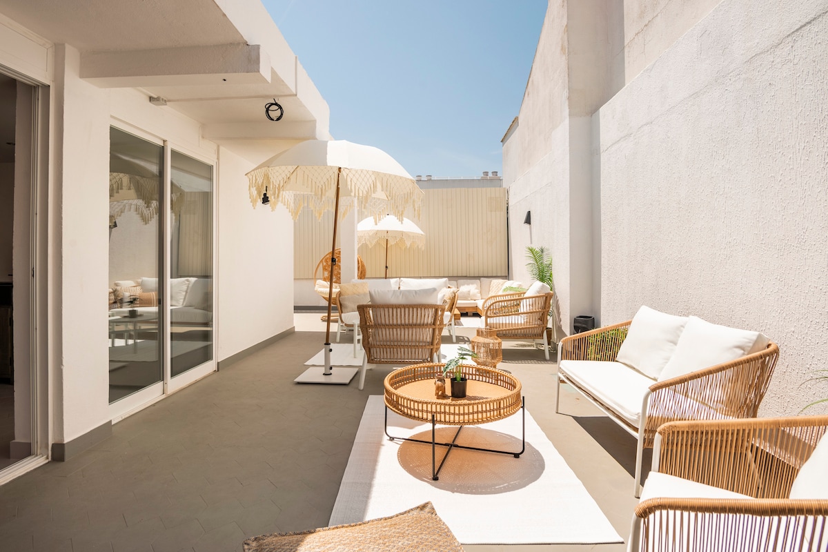A spacious terrace area is illuminated by natural light, featuring stylish rattan seating, a central round table, and a sun umbrella. The clean surfaces and minimalist design create an inviting atmosphere for relaxation and socializing.