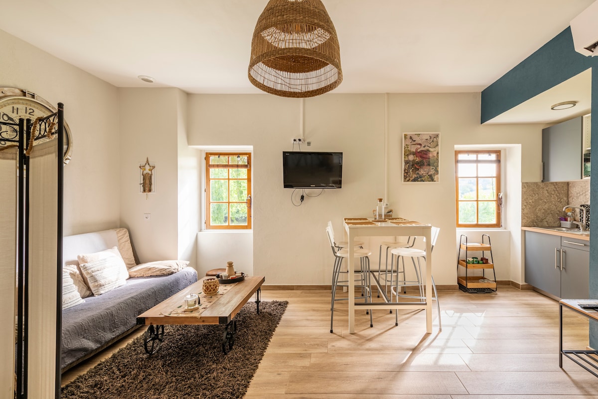 A modern studio is presented, featuring a cozy seating area with a sofa and coffee table. A dining table with high chairs sits adjacent to a kitchenette. Natural light filters through two windows, illuminating the warm tones of the decor and wooden accents.