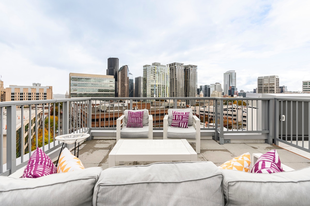 The rooftop terrace is equipped with comfortable seating, including a plush sofa and outdoor chairs. A coffee table is positioned in the center. The urban skyline features a mix of modern buildings, providing a scenic backdrop under an overcast sky.