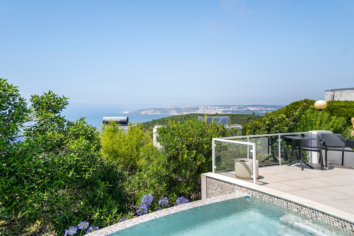 A serene outdoor space features a private pool surrounded by lush greenery and vibrant flowers. A barbecue area is visible, with expansive views of the ocean and distant coastlines under a clear blue sky.
