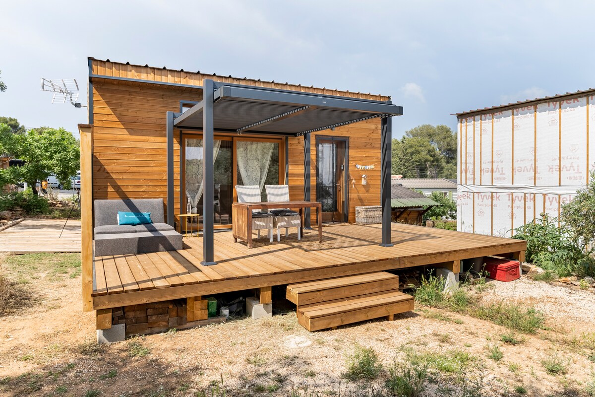 A wooden chalet exterior is shown, featuring a spacious deck with a modern awning. Two lounge chairs and a wooden table are positioned for outdoor relaxation. Steps lead up to the entrance, framed by a lush green landscape and a serene atmosphere.