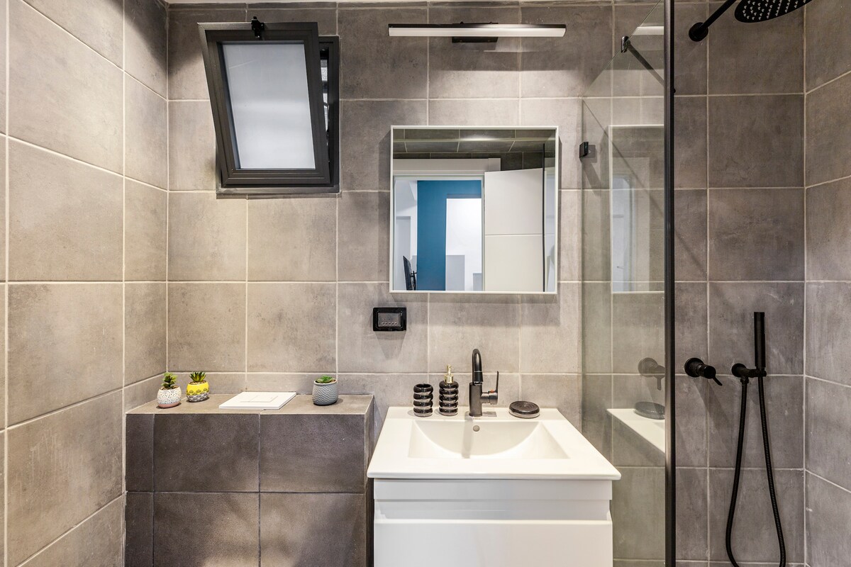 A modern bathroom is featured, showcasing a sleek white vanity with a basin and a large wall-mounted mirror. The light grey tiled walls are complemented by a glass shower enclosure. A window offers natural light, while minimal decor adds a subtle touch.