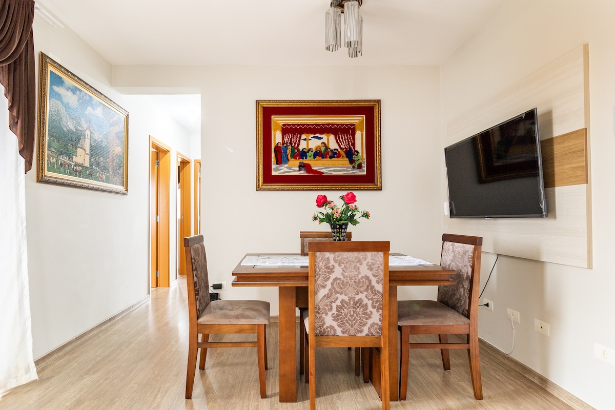 A dining area features a wooden table surrounded by four upholstered chairs. A vase with roses sits at the center, and a large wall-mounted television is visible nearby. Framed artwork adorns the walls, adding a touch of color to the serene space.