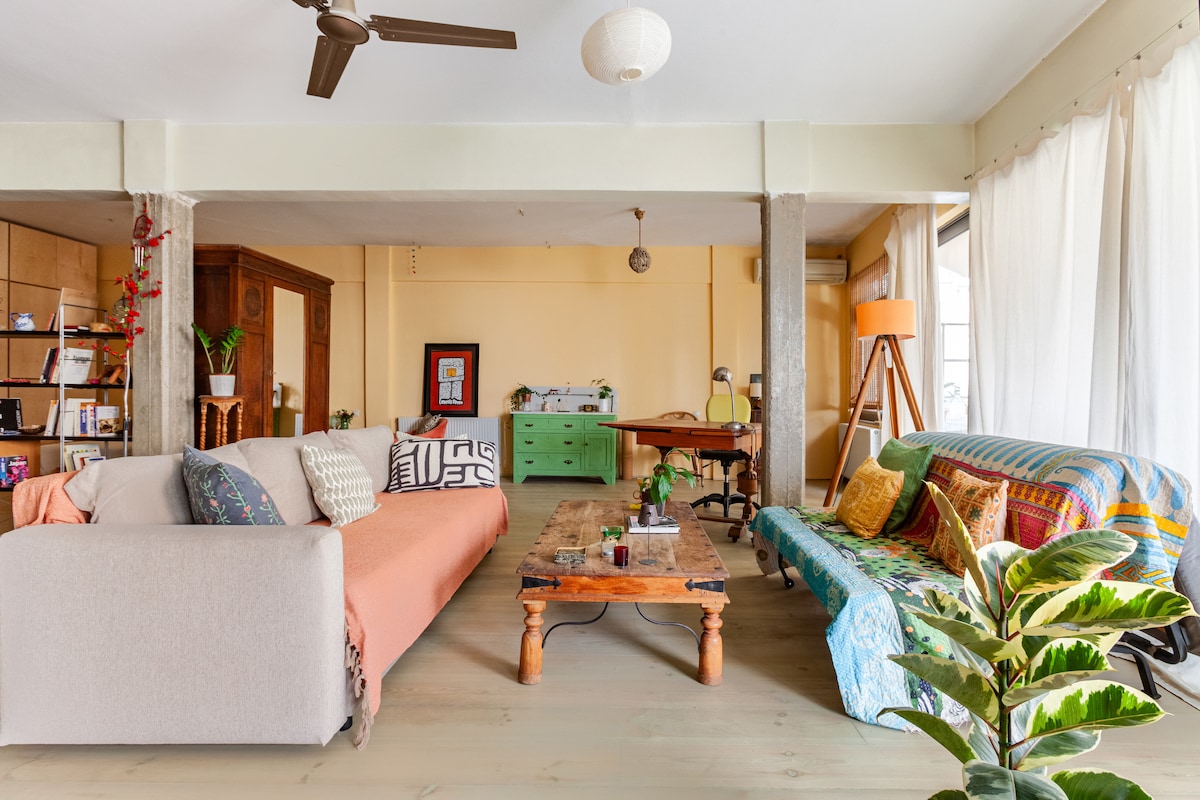 An airy living area features a mix of comfortable seating, including a pastel sofa and a colorful patterned couch. A wooden coffee table occupies the center, adorned with decorative items. Natural light floods the space through large windows, complemented by lush greenery near the floor.