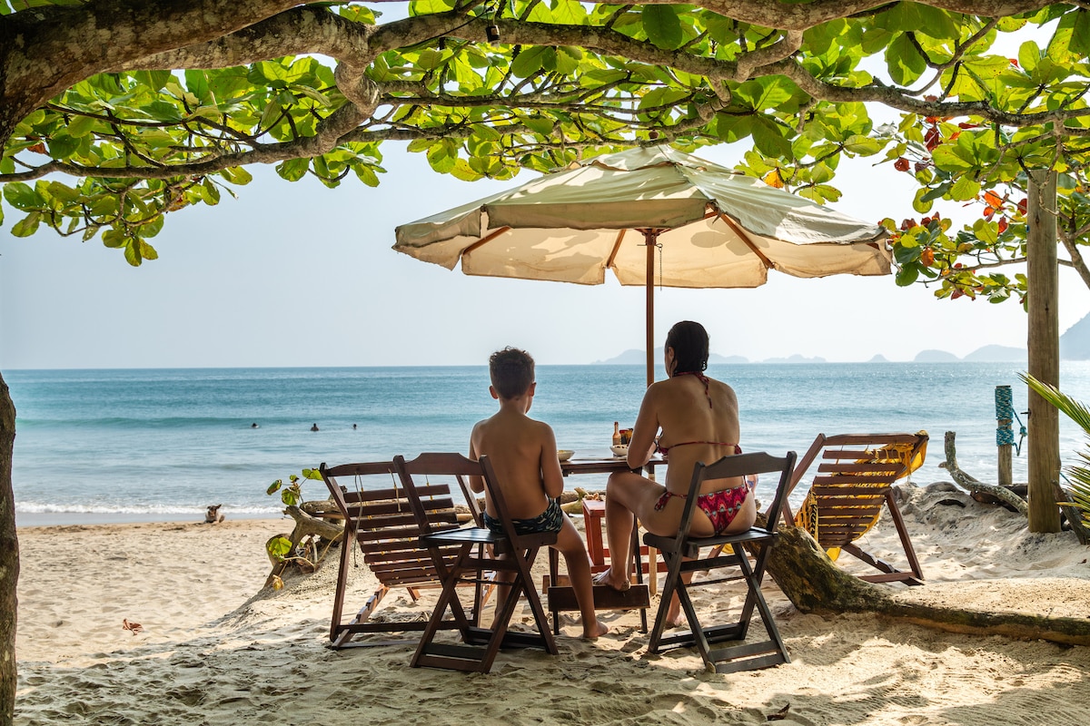 Two individuals are seated under a large umbrella, enjoying a serene beach view. Wooden deck chairs provide comfort, with the soft sand visible around them. The calm ocean stretches into the horizon, framed by lush green leaves and distant hills.