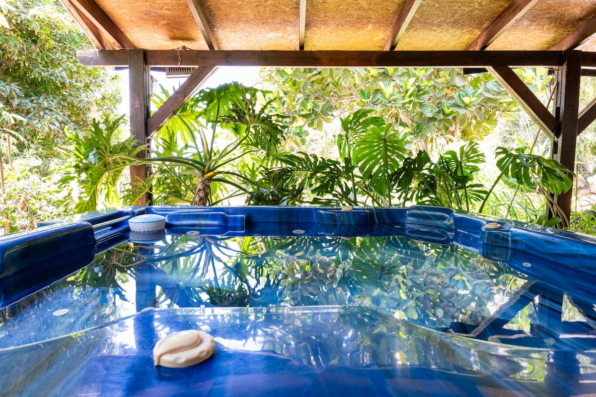 A luxurious jacuzzi is nestled under a wooden structure, surrounded by lush green plants. The water reflects the natural light, providing a serene atmosphere. Soft foliage decorates the area, enhancing the appeal of this outdoor relaxation space.