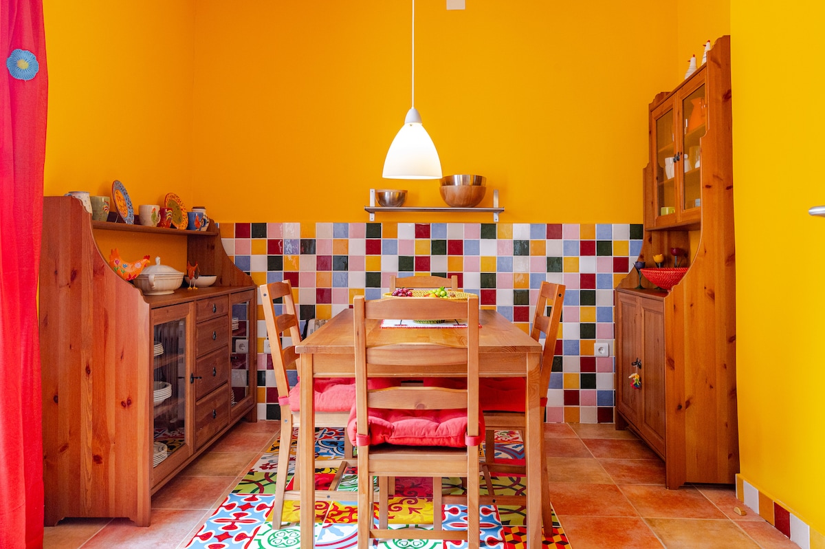A vibrant dining area is showcased, featuring a wooden table surrounded by matching chairs with red cushions. Colorful tiled walls complement the bright yellow backdrop, while a pendant light hangs above. Wooden cabinets display decorative dishware, and a textured rug adds warmth to the space.
