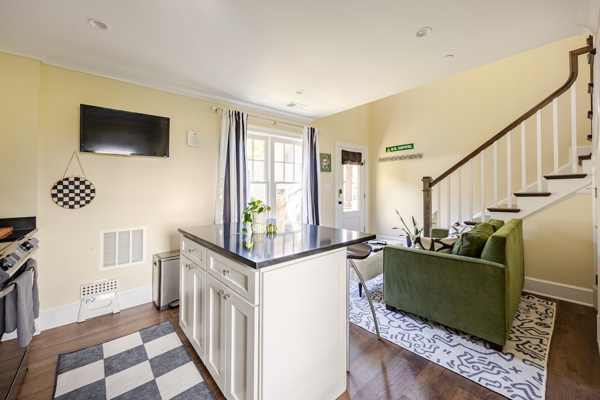 A modern kitchen area is highlighted by a central island with seating. Light yellow walls complement the dark countertops, while large windows bring in natural light. A comfortable green couch is situated nearby, creating an inviting conversation space. Decorative accents add subtle charm.
