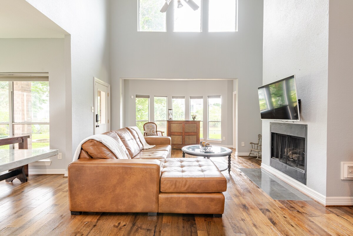 A spacious living area features a light brown leather sectional sofa and a round coffee table, positioned near a fireplace. Large windows invite natural light, highlighting the wooden floor and creating a bright atmosphere. An adjacent room is visible through glass doors.