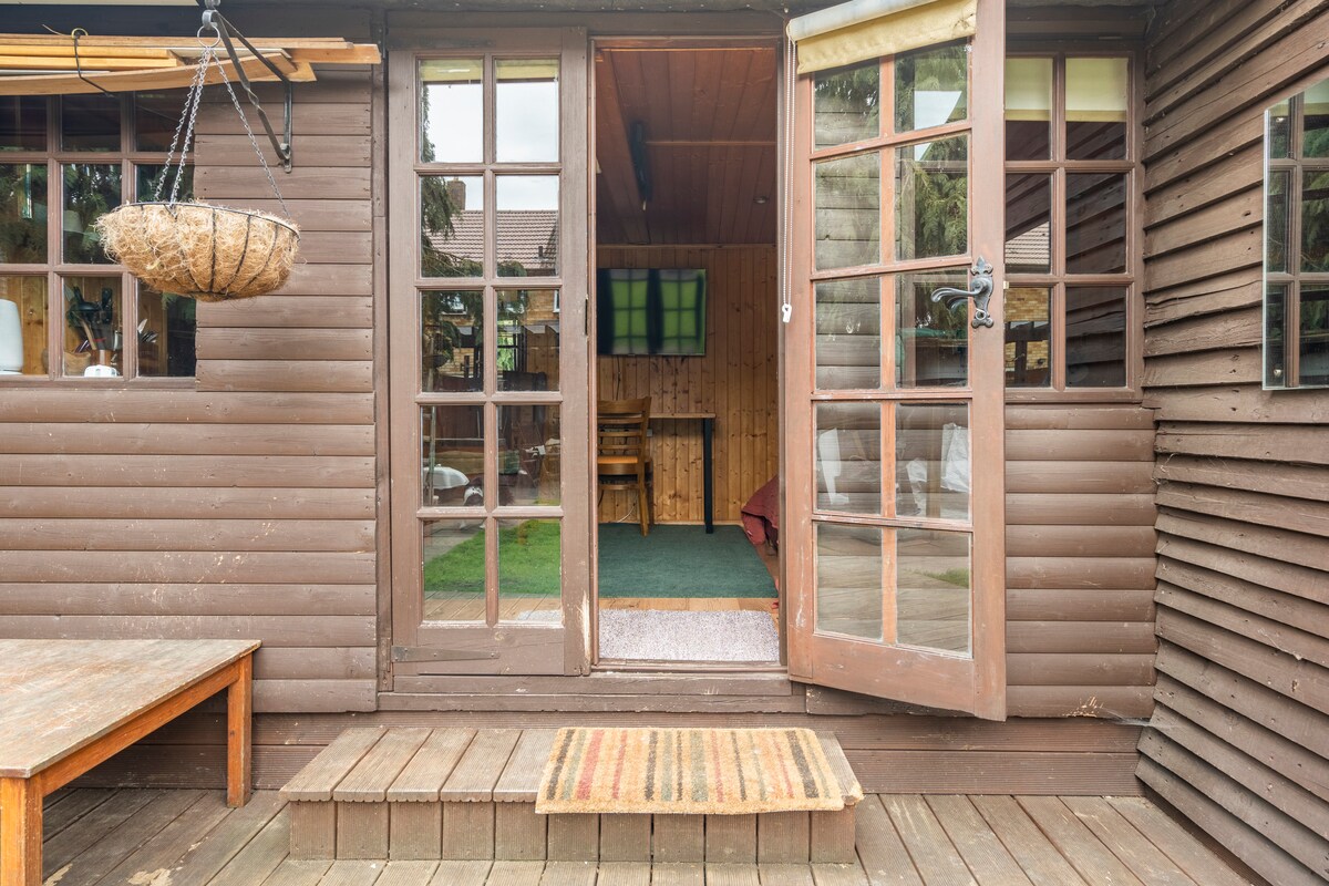 The entrance features double glass doors that open to a wooden deck, inviting natural light into the cabin. A welcome mat lies in front of the doors, while a hanging plant adds a touch of greenery. The interior is visible, showcasing a casual seating area.