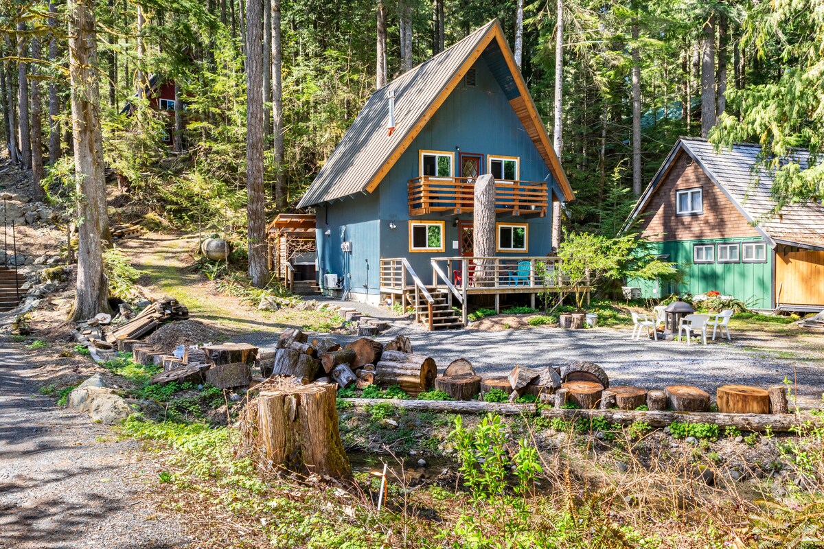 A classic A-frame cabin is nestled in a forested area, featuring a blue exterior with wooden accents. A spacious deck with railings provides access to the cabin, surrounded by tall trees and a gravel pathway. Seating areas with logs are arranged in front.