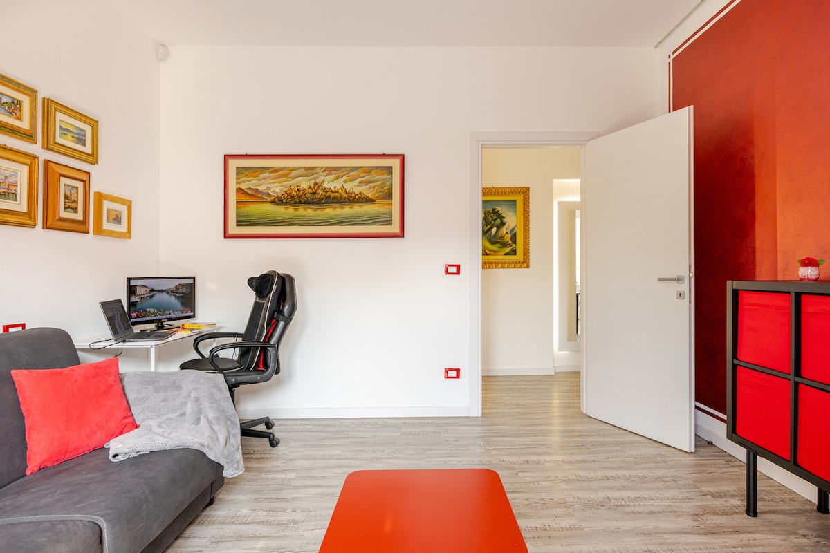 A modern living area presents a comfortable gray sofa adorned with red cushions. A bright red coffee table sits in front. An art piece displays vibrant colors, while a small work desk and chair are positioned nearby. Light wood flooring complements the light-filled space.
