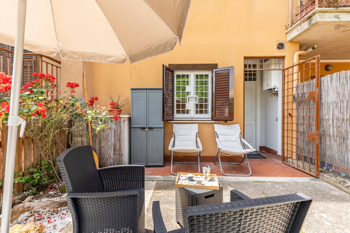 An outdoor sitting area is highlighted, featuring two lounge chairs positioned next to a small table. Windows are open, allowing natural light to enhance the space. A parasol provides shade, and vibrant flowers create a pleasant backdrop.