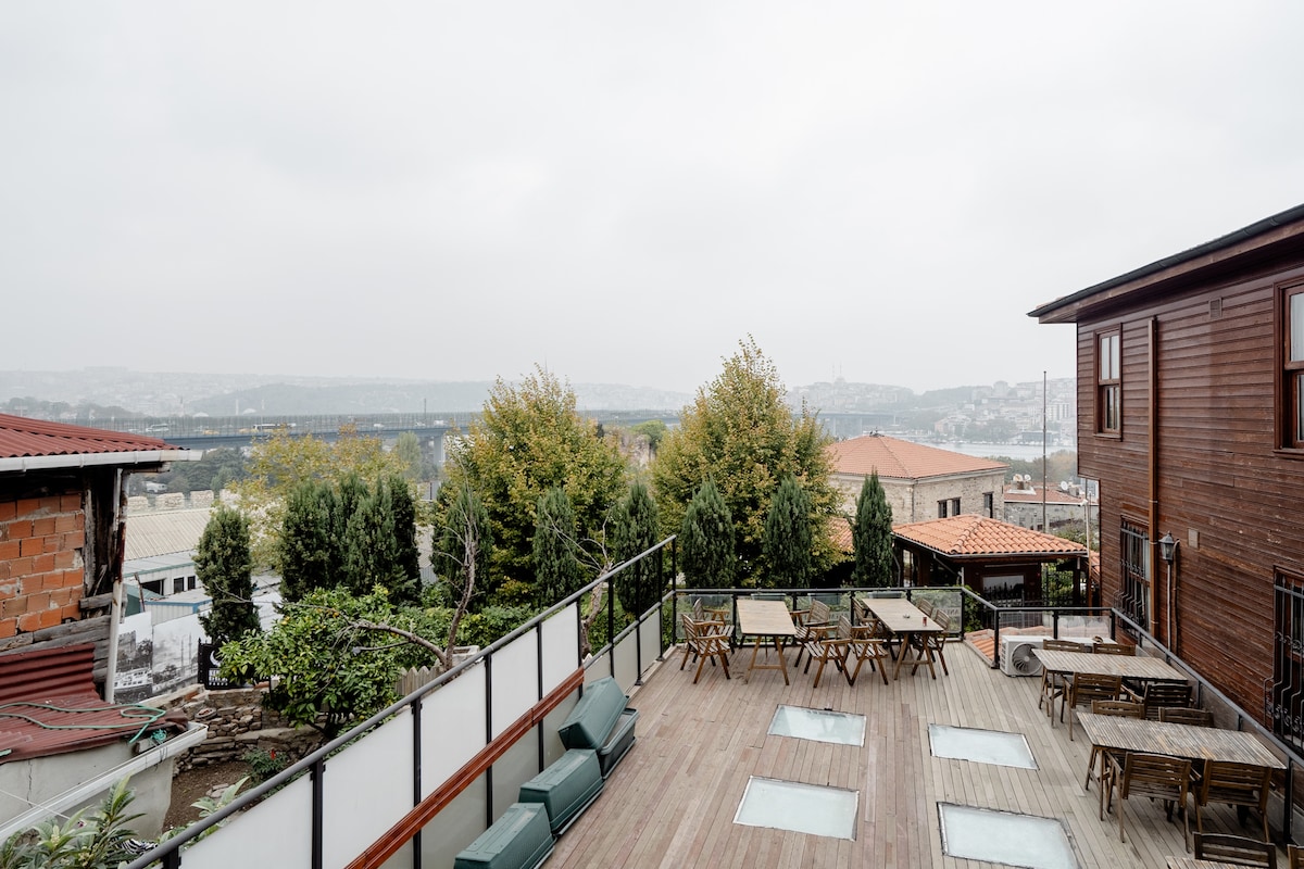 An outdoor terrace is presented, featuring several wooden tables and benches arranged for dining. Lush greenery is visible, along with a distant view of a bridge. The wooden structure of the building complements the natural setting.