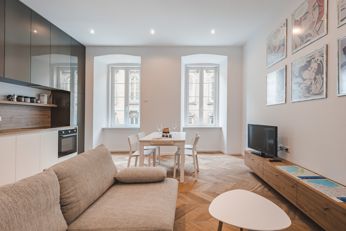 A modern living space is highlighted by large windows allowing natural light. The open kitchen features sleek cabinets and a compact dining area with a table for four. A comfortable sofa is positioned nearby, alongside a television and decorative wall art.