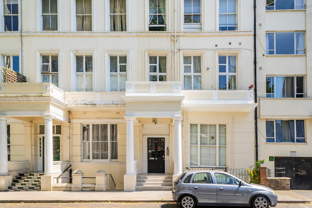 The elegant building features a classic facade with large windows allowing natural light to enter. A welcoming entrance is accentuated by white columns and steps. A parked car is visible in front, adding a sense of urban life to the scene.