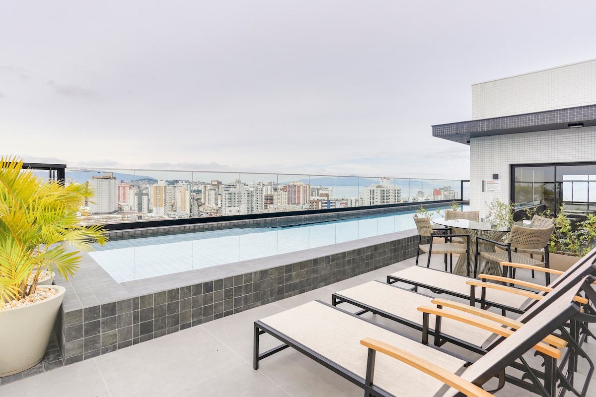 A rooftop pool area is showcased, featuring a clear pool that reflects the skyline. Comfortable lounge chairs are arranged around the pool, complemented by a few tropical plants in elegant planters. The space offers expansive views of the city and ocean in the background.