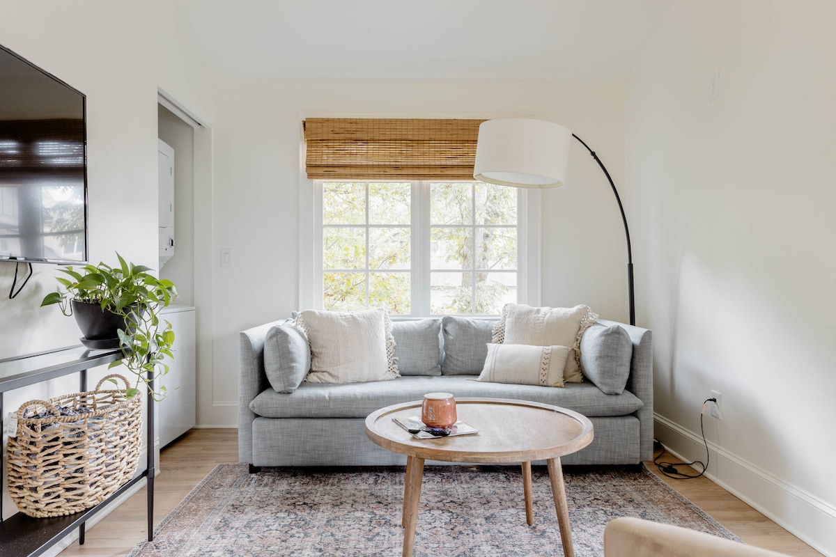 A bright living area is presented with a soft gray sofa adorned with decorative pillows. A round wooden table occupies the center, accompanied by a plant in a woven basket. Natural light filters through the large windows, enhancing the inviting ambiance.