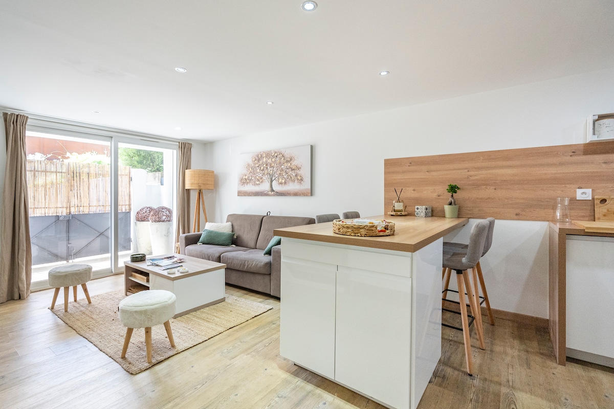 A contemporary open-plan living area features a light grey sofa and wooden accents. Natural light enters through sliding glass doors leading to an outdoor space. A dining table with stools is positioned near a wooden kitchen countertop, enhancing the inviting ambiance of the space.