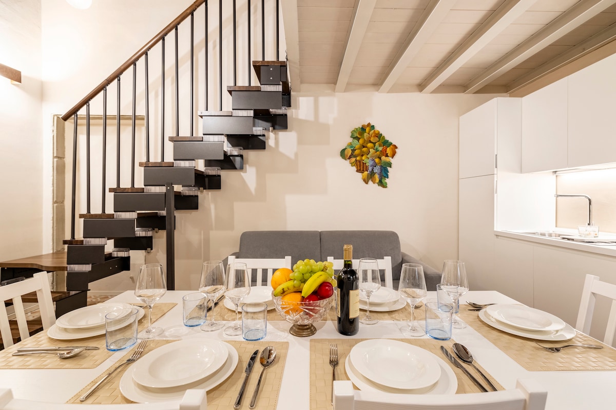 A dining area features a large table set for six, complete with plates, glasses, and a fruit bowl in the center. A modern kitchen is visible in the background, and a staircase leads to the upper level. Soft lighting highlights the white walls and wooden beams.